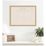 Beige linen fabric pinboard designed like a vintage postcard with gold frame mounted on a white wall above a white surface holding a lamp, photo frame, and potted plant.