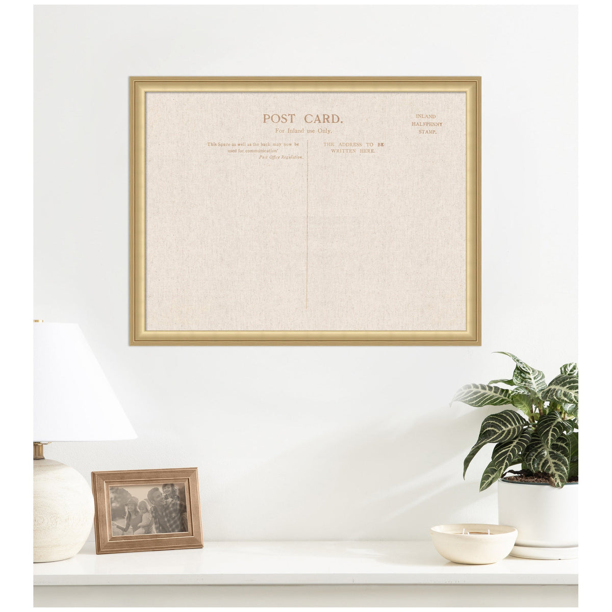 Beige linen fabric pinboard designed like a vintage postcard with gold frame mounted on a white wall above a white surface holding a lamp, photo frame, and potted plant.