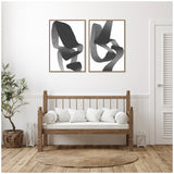 Two framed abstract black and white canvas prints with flowing brushstroke shapes hung above a wooden bench with white cushions on a white wall, beside a plant and a round woven rug.