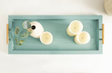 Rectangular teal tray with raised edges and gold handles, holding a small white vase with green leaves and three cream-colored candles.
