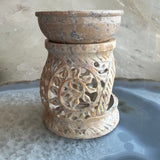 Carved beige and brown soapstone essential oil burner with floral cutout pattern and rounded bowl on top, standing on a gray and blue marbled surface.