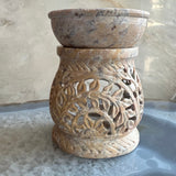 Carved soapstone essential oil burner with intricate floral and leaf patterns in warm beige and brown tones, shown upright on a reflective surface against a textured wall.