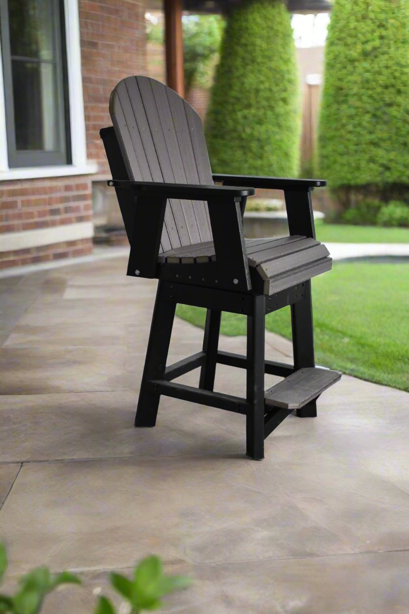 Tall Adirondack chair with a black frame and dark gray slatted seat and backrest, placed on a smooth concrete patio near a brick wall and green shrubs.