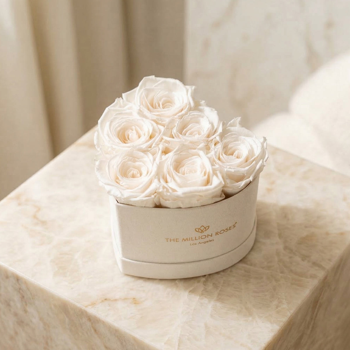 Heart-shaped beige box holding six white roses with soft petal texture, placed on a cream-colored marble surface in natural light.