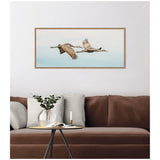 Framed canvas wall art above a brown couch shows two sandhill cranes flying side by side against a pale blue sky with soft clouds.