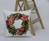 Square white throw pillow featuring a colorful Christmas wreath with red berries, poinsettias, pine cones, and green foliage, placed next to a wooden step ladder.