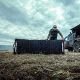 Man setting up a large black foldable Dark Energy solar panel on dry grass beside a rugged off-road vehicle under a cloudy sky
