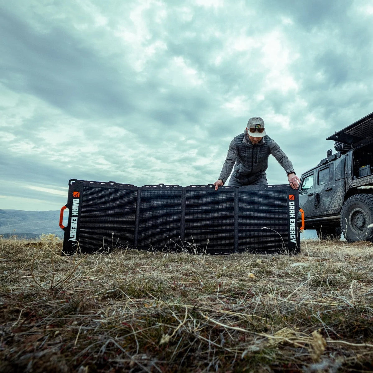 Man setting up a large black foldable Dark Energy solar panel on dry grass beside a rugged off-road vehicle under a cloudy sky