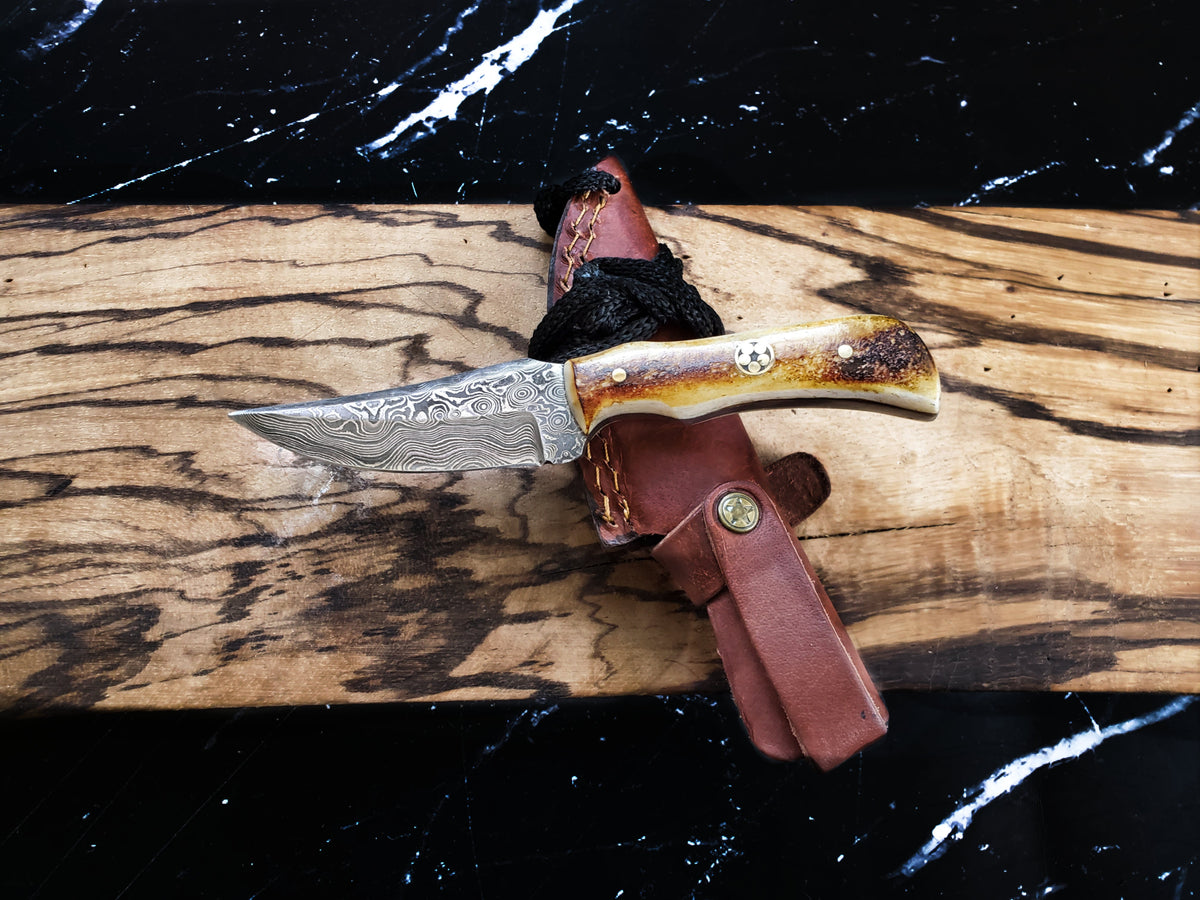 Hand forged Damascus Knife Neck Knife TD-178 Burnt Bone resting on a wooden surface with its leather sheath.