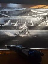 Black and white etched artwork depicting a hockey player skating on ice with a large arena labeled 'Steel Energy Centre' and starry sky in the background, with a Dremel tool in foreground.