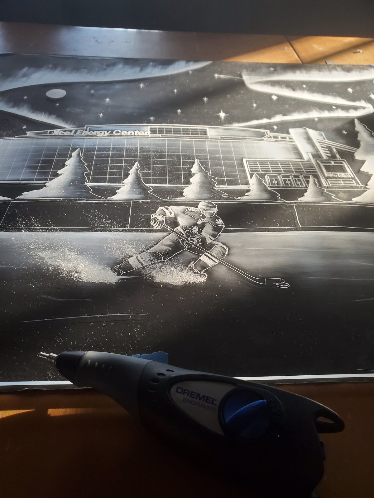 Black and white etched artwork depicting a hockey player skating on ice with a large arena labeled 'Steel Energy Centre' and starry sky in the background, with a Dremel tool in foreground.