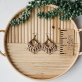 Pair of beige macrame fringe earrings with diamond-shaped wooden frames, displayed on a round wooden tray with a small succulent and a measurement card.