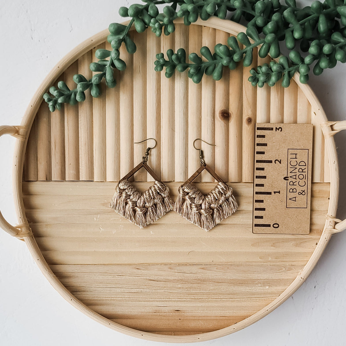 Pair of beige macrame fringe earrings with diamond-shaped wooden frames, displayed on a round wooden tray with a small succulent and a measurement card.