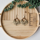 Pair of beige macrame fringe earrings with bronze triangular frames placed on a round wooden tray next to a 3-inch ruler and green faux succulent branches.