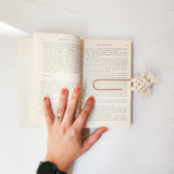 Hand with coral nail polish and rings holding open a book, edged by a cream-colored macrame bookmark with tassels on a white textured surface.