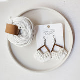 Pair of macrame fringe earrings with wooden triangular frames and white woven fringes, displayed on a white card next to a coil of thick white cord on a round plate.