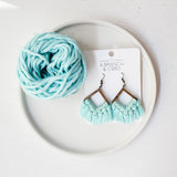 Macrame fringe earrings with light turquoise cotton cord and a wooden frame, displayed on a white card inside a white shallow dish with a ball of matching colored cord.