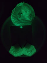Glowing green macrame fringe earrings suspended on a dark backing card, showing detailed knotting and texture in low light.