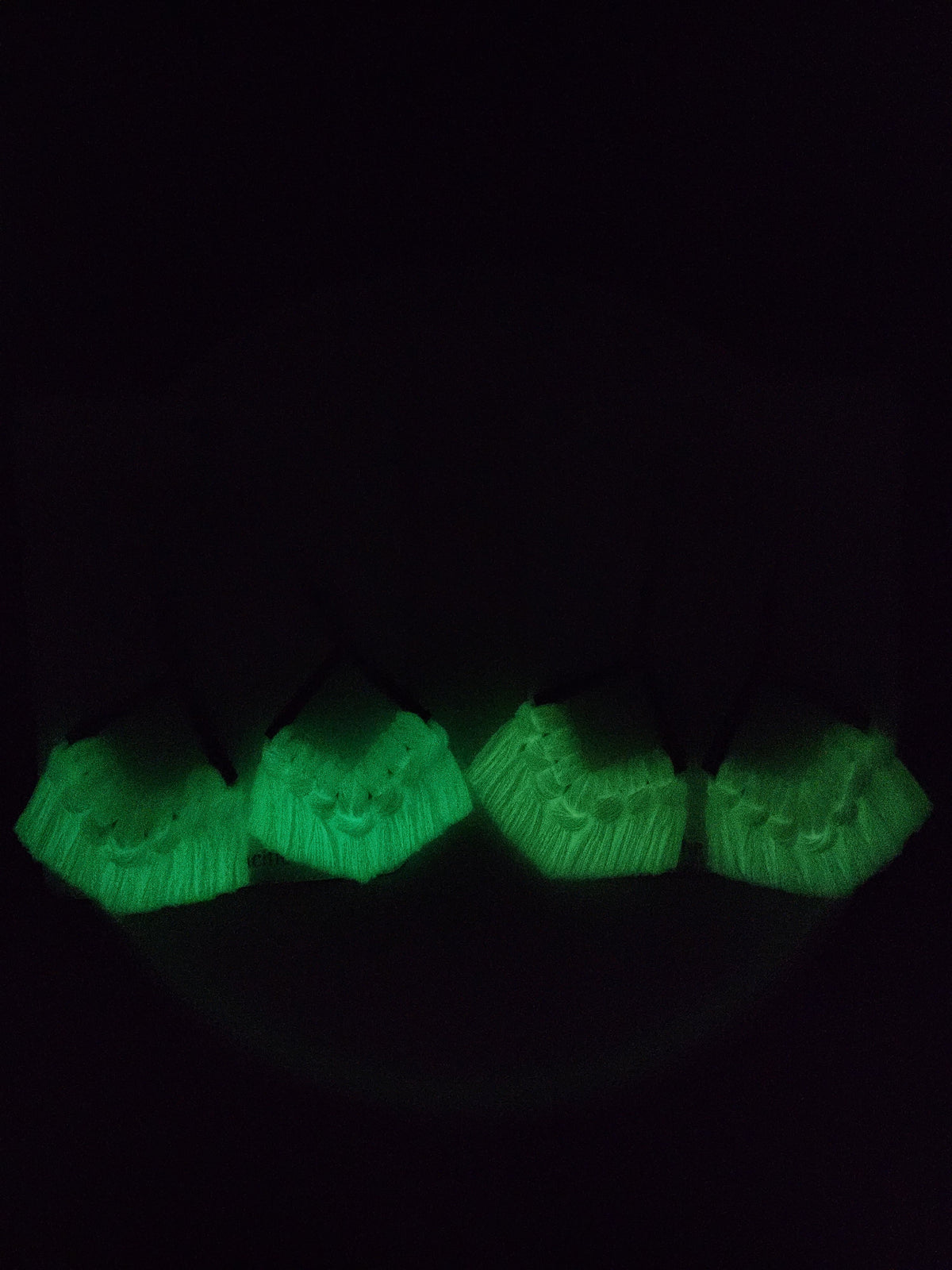 Four pairs of macrame earrings with glowing green fringes, arranged in a row against a dark background, highlighting their luminous texture and fringe details.