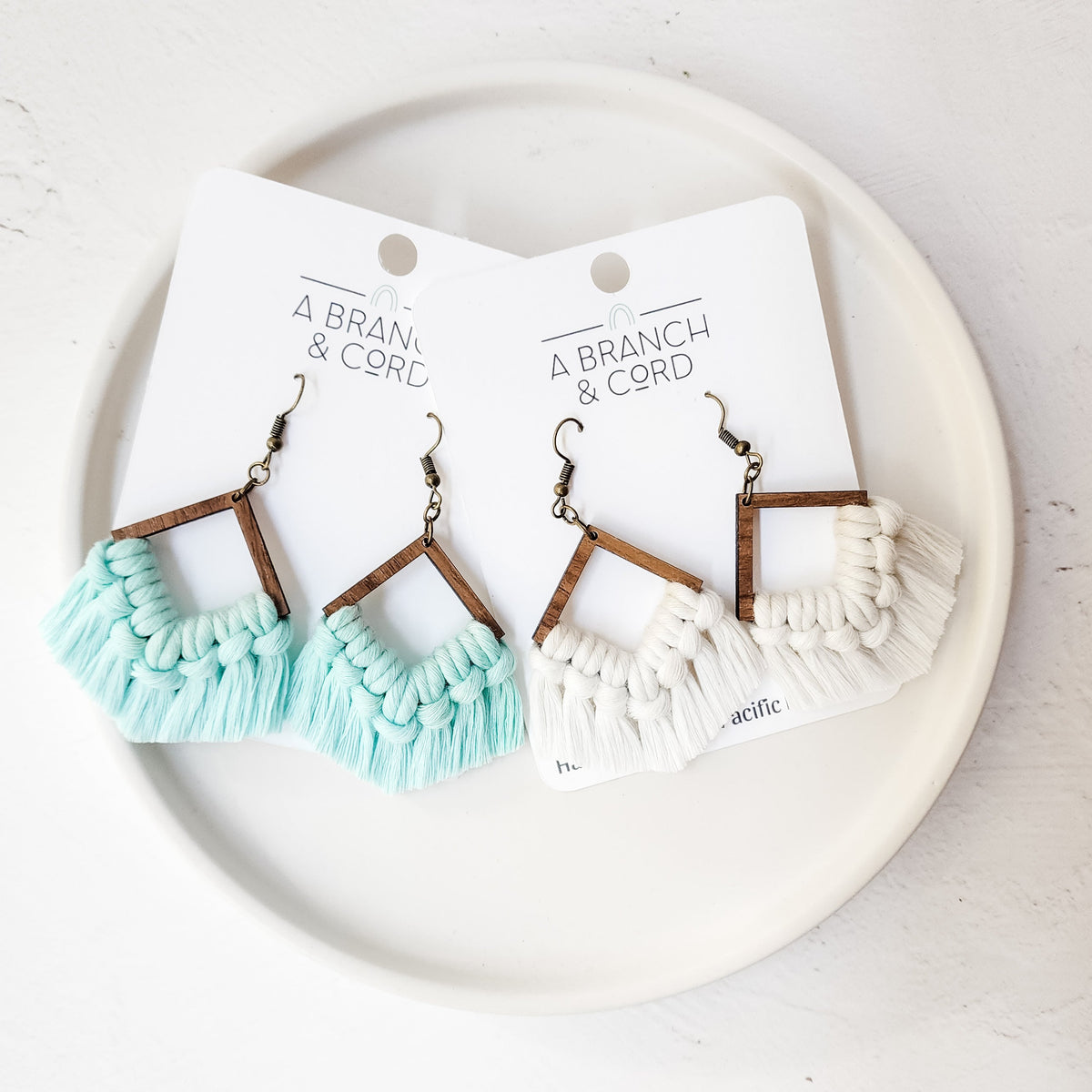 Blue glow-in-the-dark macrame fringe earrings hanging from a branch with soft natural background