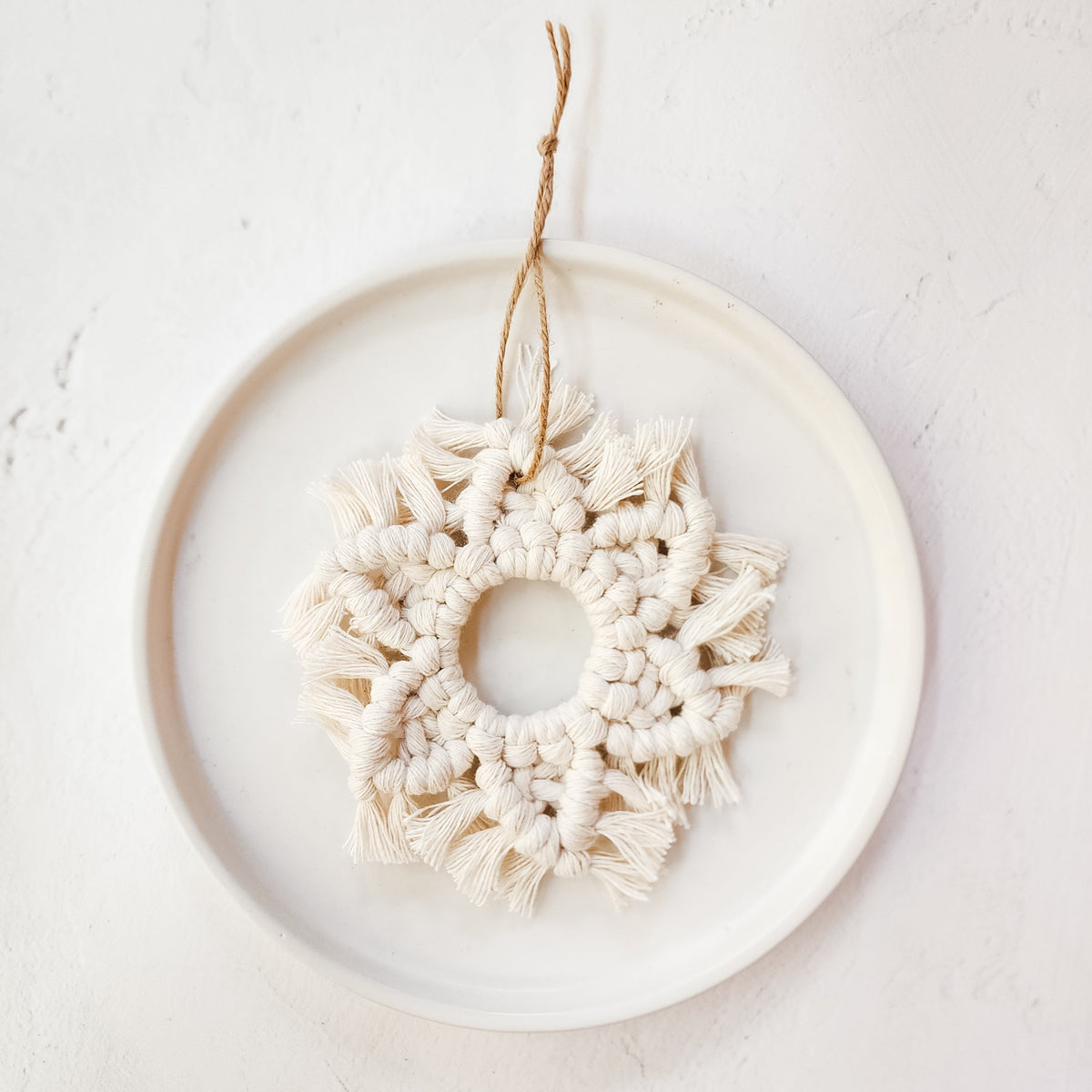 Cream-colored macrame snowflake ornament with fringed edges hanging on a white ceramic plate against a textured white wall.