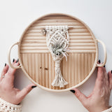 Hand holding round bamboo tray with horizontal slats, featuring a small cream-colored macrame hanger holding a green air plant at center.