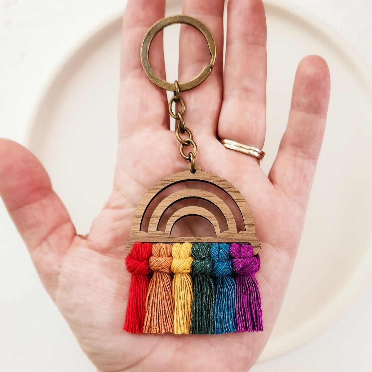 Hand holding a wooden rainbow keychain with six colorful yarn tassels in red, orange, yellow, green, blue, and purple hanging from the bottom edge