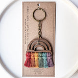 Wooden rainbow keychain with five arches, each with hanging yarn tassels in red, orange, yellow, green, and purple tones, attached to a metal key ring on a brown card.