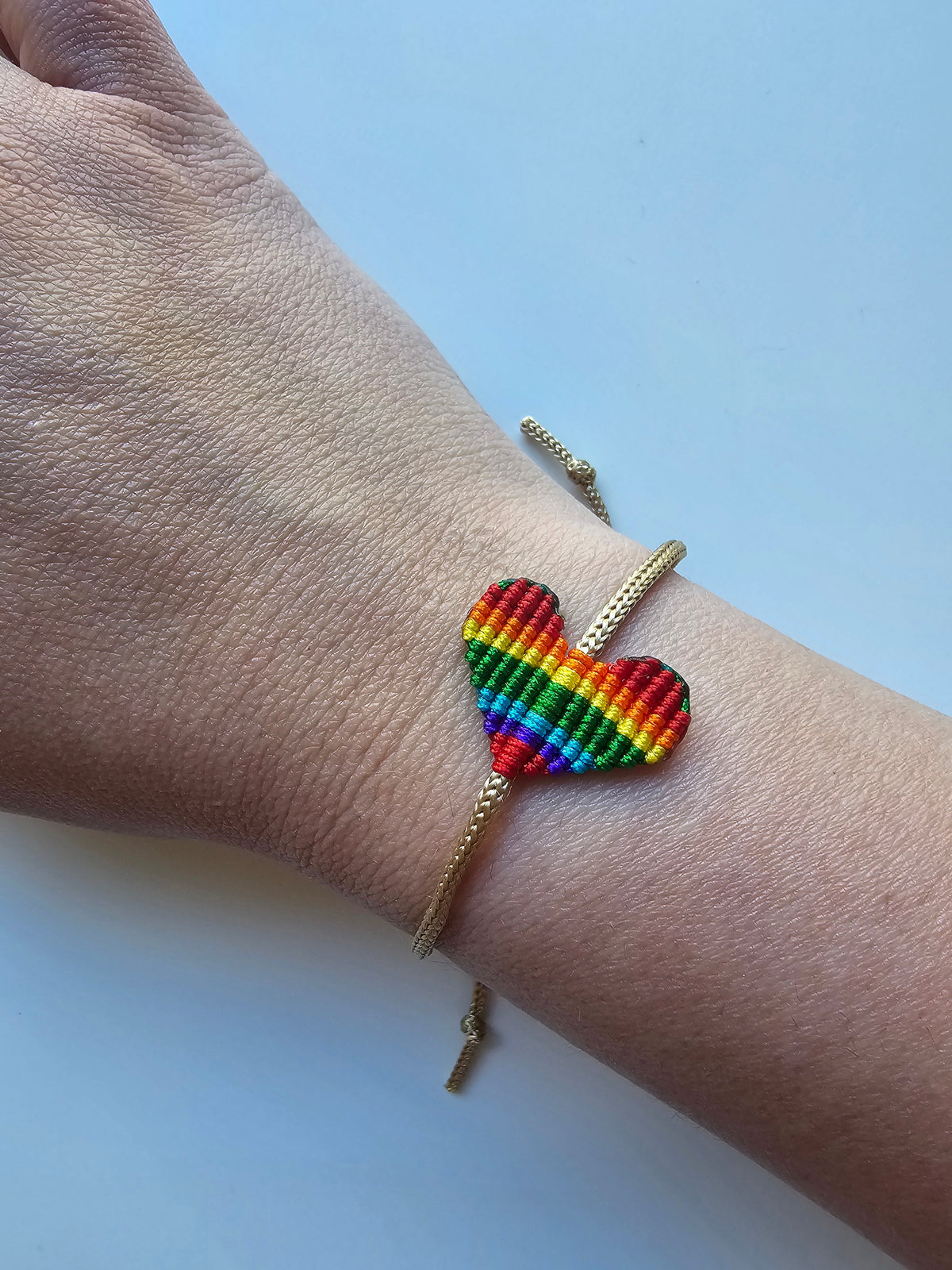Wrist wearing a macrame bracelet with a heart-shaped centerpiece woven in rainbow pride colors and adjustable gold cord.