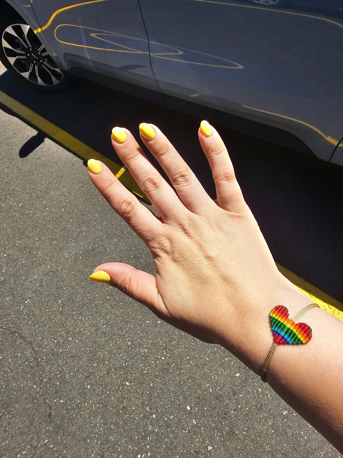 Hand with bright yellow nail polish wearing a rainbow-striped heart bracelet, held over asphalt near a gray car with a reflection on its side.
