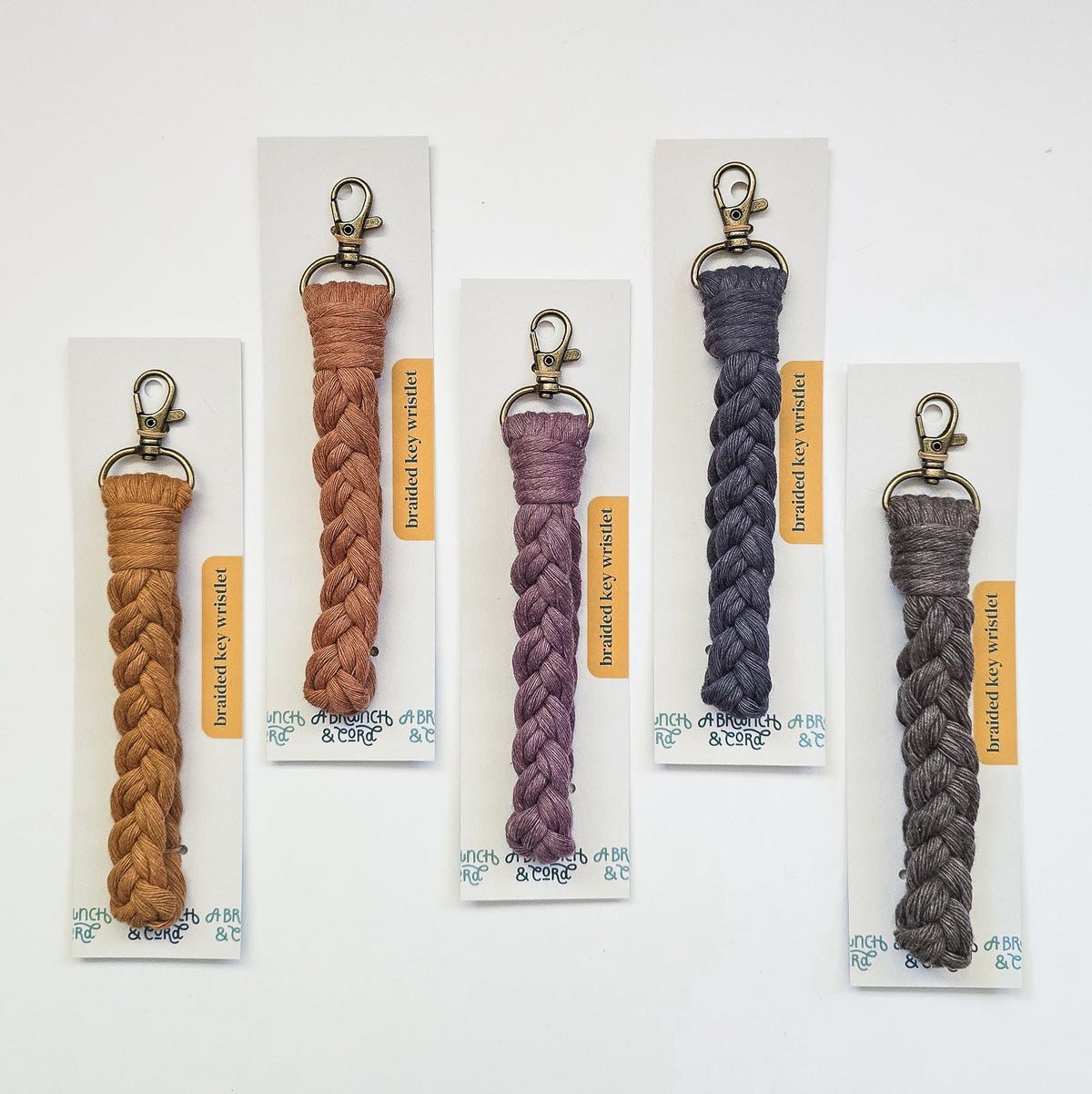 Five braided macrame key wristlets in varying neutral tones from mustard to gray, each with a metal clasp and attached to white cards with orange labels.
