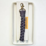 Purple-gray macrame braided key wristlet with antique brass lobster clasp, displayed on white packaging with orange label on an off-white rectangular dish.