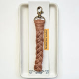 Braided neutral-toned macrame key wristlet with a brass lobster clasp, displayed on a rectangular white ceramic tray with a product card beneath.