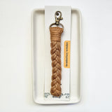 Light brown macrame braided key wristlet with a gold clasp, displayed on a white card resting on a rectangular white ceramic tray.