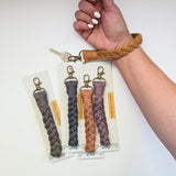 Braided macrame key wristlet in tan worn on wrist with four more in gray, blue, peach, and mauve, each with a brass lobster claw clasp on display cards.