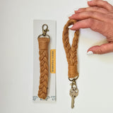 Two tan-colored macrame braided key wristlets with brass key rings and clips, one held by a hand and one attached to packaging with a label.
