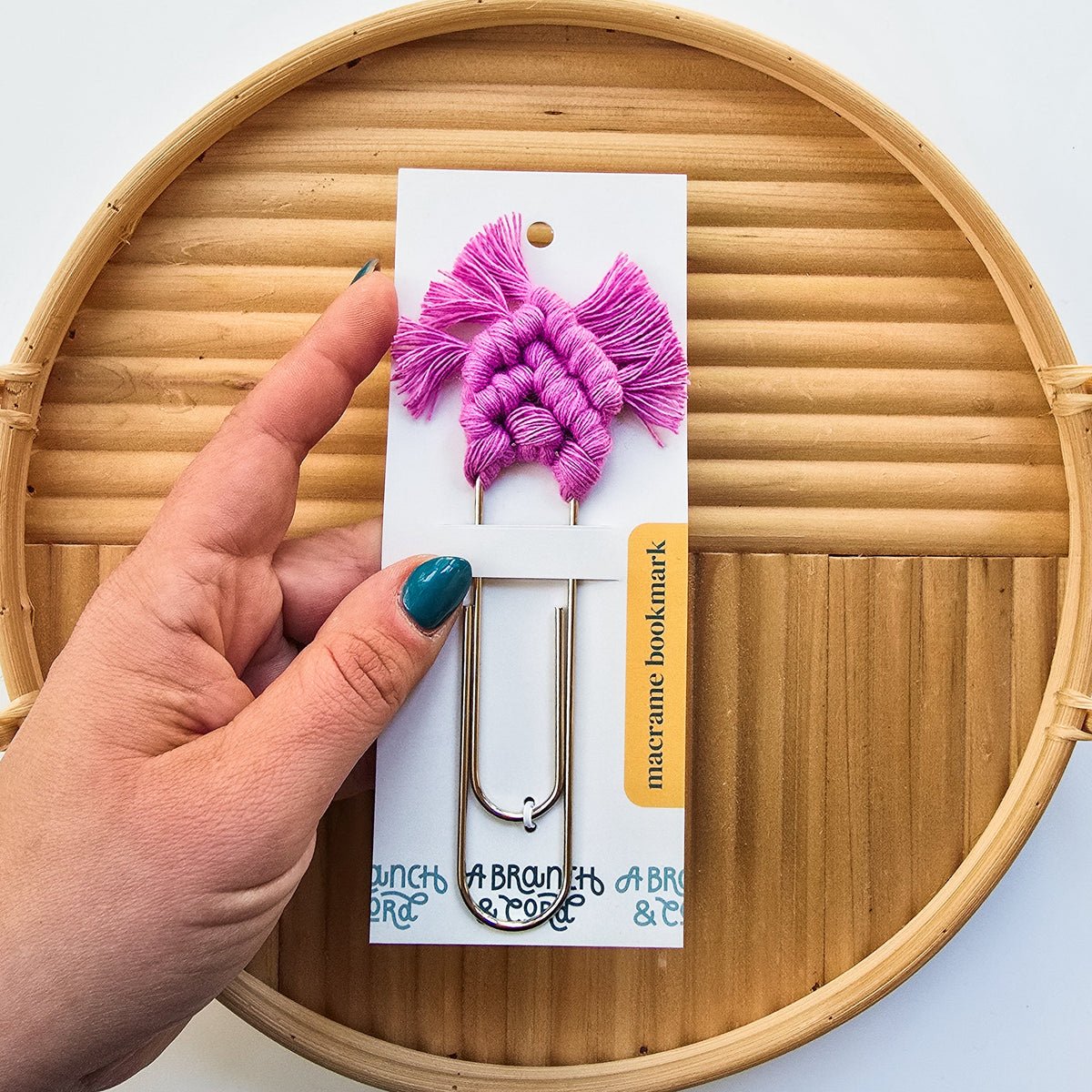 Large silver paperclip bookmark with bright pink macrame fringe held over a round woven bamboo tray, showing turquoise-painted fingernails.