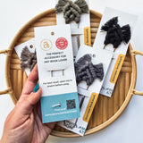 Hand holding a neutral-toned macrame bookmark on white card with metal clip, displayed over a round bamboo tray holding four similar bookmarks in black and gray tones.