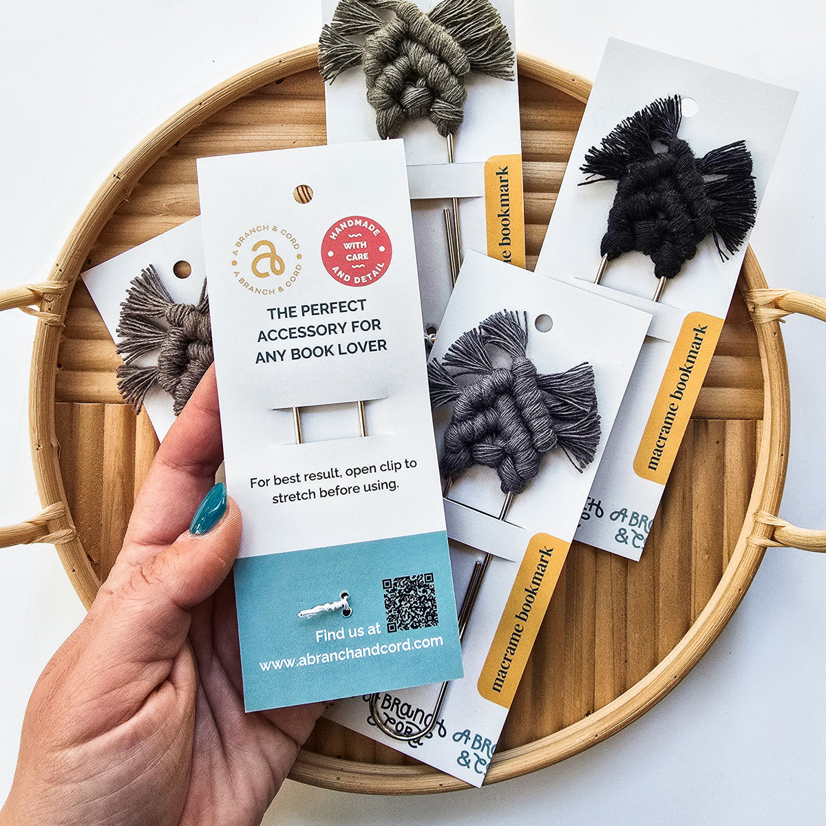 Hand holding a neutral-toned macrame bookmark on white card with metal clip, displayed over a round bamboo tray holding four similar bookmarks in black and gray tones.