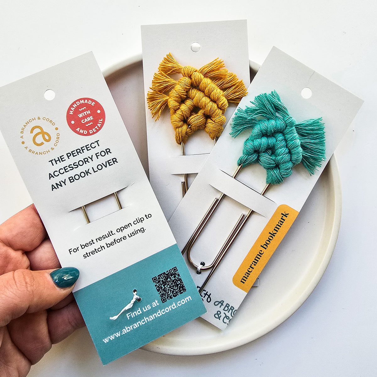 Two handmade macrame bookmarks in mustard yellow and teal, each with fringe, displayed on white cards over a white ceramic plate with a hand holding one card.