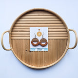 Round wooden tray with horizontal ridges on the top half and smooth wood with knots on the bottom, holding macrame knotted earrings with brown hoops and burnt orange details on a white card.