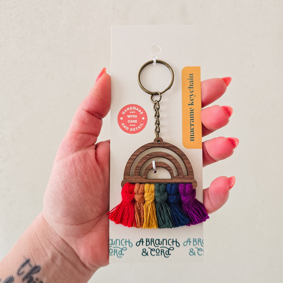 Hand holding a macrame keychain with a wooden rainbow shape featuring stripes and colorful tassels in red, orange, yellow, green, blue, and purple, attached to a metal ring.