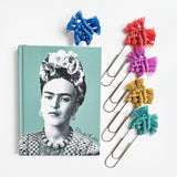 Four large silver paperclips each adorned with a different bright macrame knot in blue, red, purple, yellow, and teal, clipped to a teal book with a black-and-white portrait of Frida Kahlo.