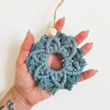 Hand holding a circular blue macrame snowflake ornament with fringed edges and a wooden bead on top, suspended by a natural twine loop.