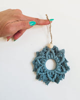 Hand holding a blue macrame snowflake ornament with a wooden bead and a jute hanging loop against a white background.