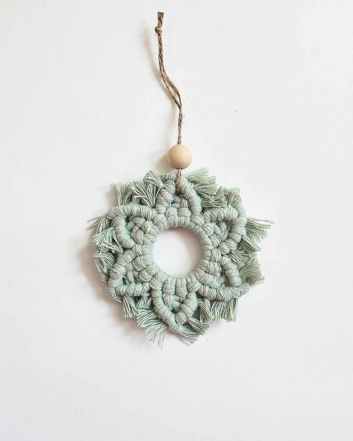 Light green macrame snowflake ornament with knotted pattern and fringed edges, hanging from twine with a single wooden bead at the top.