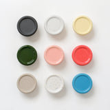 A grid of nine round coat hooks in solid matte and glossy finishes arranged on a white surface, featuring colors black, white speckled, cream, dark green, pink, red, beige, white with confetti, and blue.