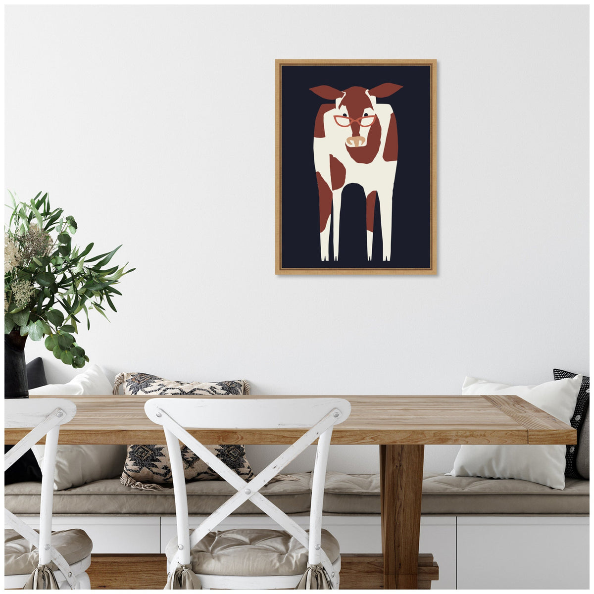 Framed minimalist canvas of a brown and white cow on a dark navy blue background hanging above a wooden dining table with white cross-back chairs and a vase of greenery.