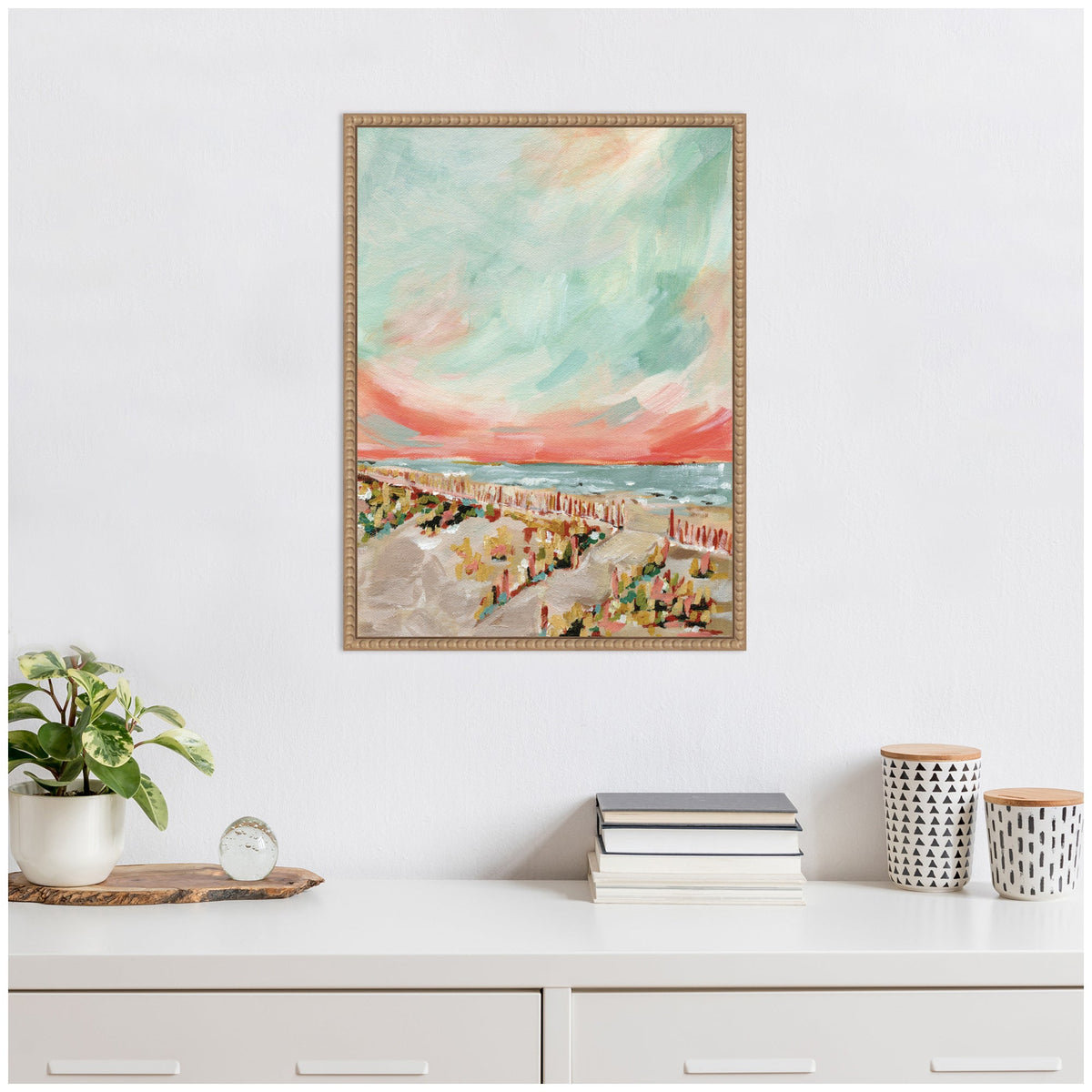 Framed Sylvie Landscape 03 Beach canvas painting by Annie Quigley featuring soft coral sky and muted teal ocean with beach scene below, hanging above white shelf with books and decor.