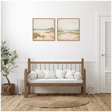 Two square framed canvas paintings above a wooden bench seat with white cushions, featuring soft pastel landscapes with sandy tones and blue skies in a bright, minimalist room.